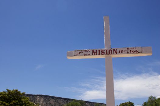 A white wooden cross/ crucifix denoting information about a Mission ahainst a blue sky, with mountains in the background with copy space.