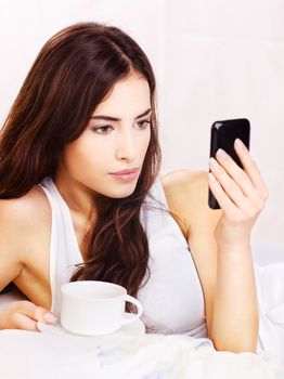 Pretty woman making phone call and holding cup of coffee in bed