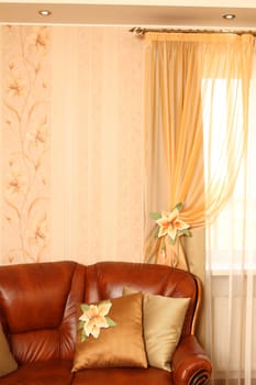 Brown leather sofa with beautiful pillows, a row a window with a curtain. Vertical shot. Close up. The embroidery isn't a handiwork, this repetition of drawing on wall-paper.