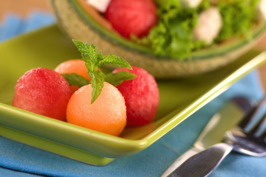 Refreshing cantaloupe and watermelon balls garnished with mint with a fresh salad in the back (Very Shallow Depth of Field, Focus on the front of the cantaloupe melon ball)