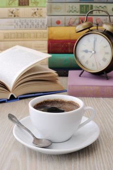 A cup of coffee on the table against the background of an open book with a clock and a stack of books