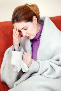 A young sick woman with a cup of tea in her hand. She hold her head in pain