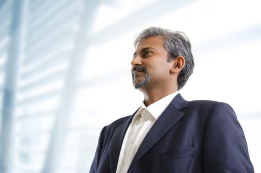 Mature Asian Indian businessman looking away standing on office background