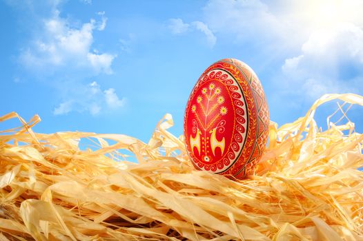 Easter egg painted with traditional ukrainian ornaments lies on yellow straw. Sunny blue sky with clouds on background