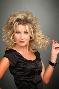 Portrait of the beautiful girl-blonde in a black dress with a magnificent hairdress. Looks in a lens, holds with a hand hair.