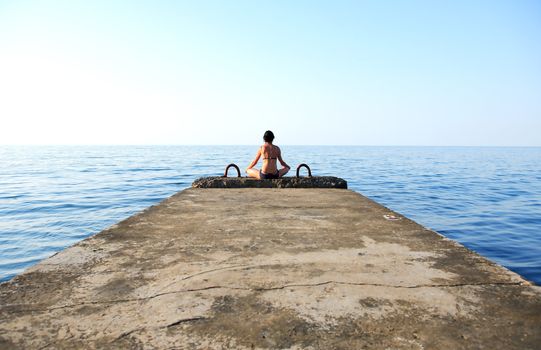 Beautiful adult woman sitting in lotus position on pier against sea