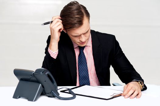 Confused young man itching his head with pen. Sitting at work desk