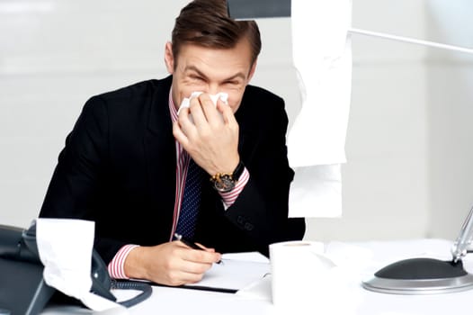 Professional sitting at desk sneezing into tissue holding pen in hand