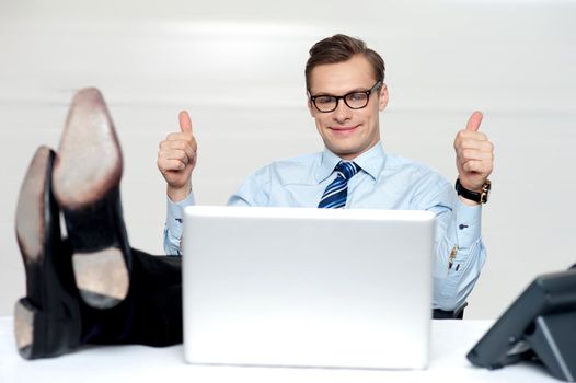 Relaxed businessman gesturing thumbs up to camera. Posing with legs on desk