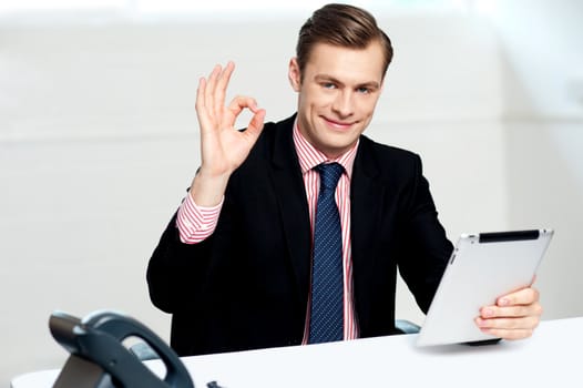 Smiling corporate male showing ok sign to camera. Holding tablet and sitting in office