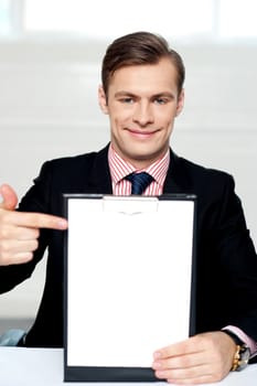 Smiling man pointing towards blank clipboard. Indoor shot