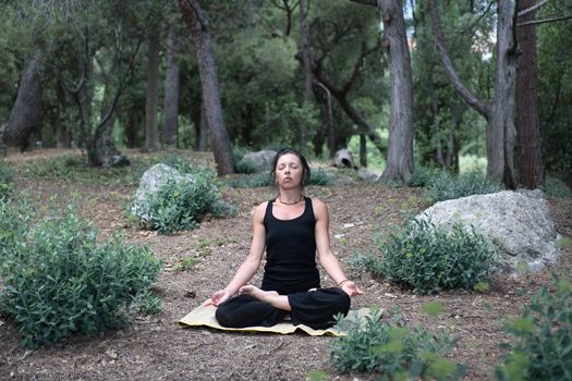 Svelte adult woman in black sitting in lotus position on forest background