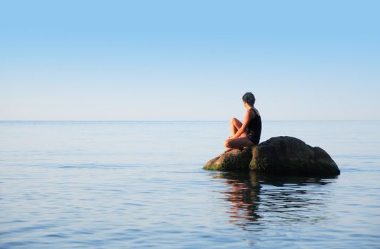 Svelte adult woman sitting on big stone in the sea and looking to horizon