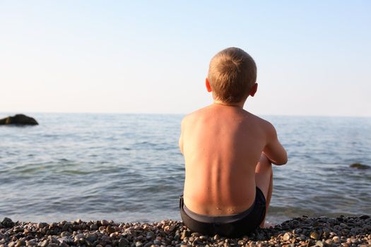Young suntan boy sitting on the beach near sea and looking to horizon
