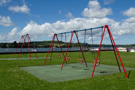 A pair of swings with red and blue frames and seats on metal chains in a park next to a stretch of water with boats backed by a blue sky with cloud.