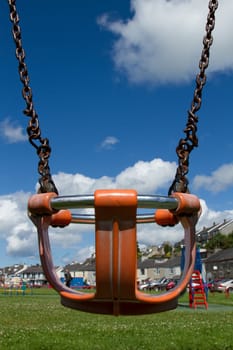 A child's safety seat with protection ring on a metal chain with green grass and a blue sky with cloud in the distance.