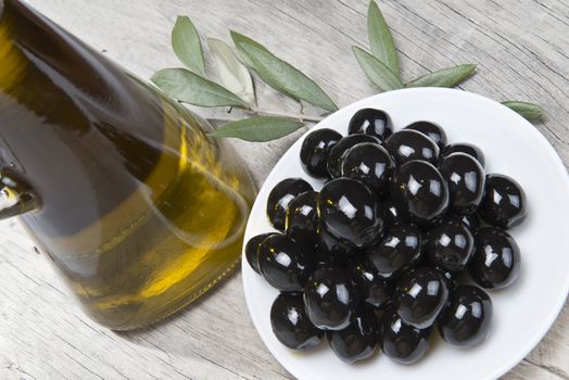 A jar with olive oil ans a plate with black olives on a wooden surface.