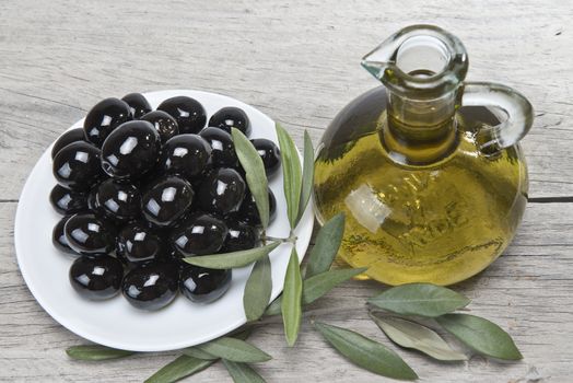 A jar with olive oil ans a plate with black olives on a wooden surface.