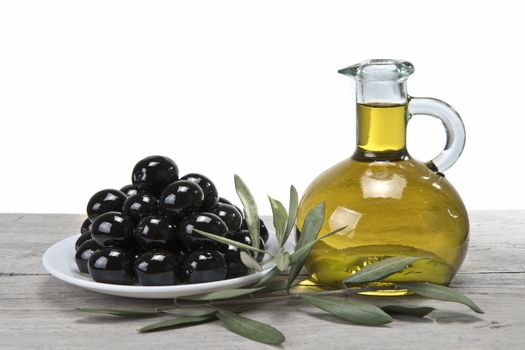 A jar with olive oil ans a plate with black olives on a wooden surface.