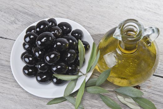 A jar with olive oil ans a plate with black olives on a wooden surface.