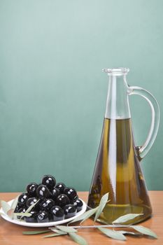 A jar with olive oil ans a plate with black olives on a wooden surface.