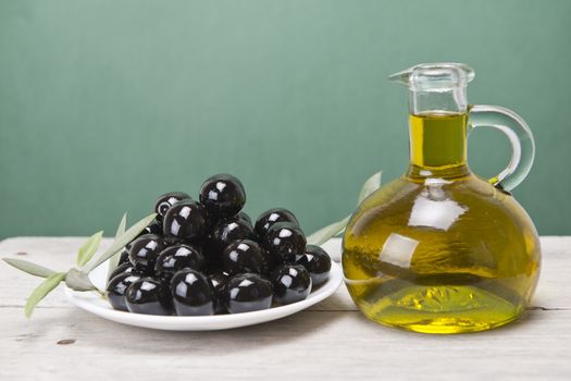 A jar with olive oil ans a plate with black olives on a wooden surface.