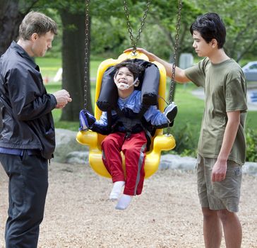 Disabled little boy swinging on special needs swing being pushed by family