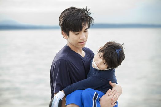 Big brother carrying disabled boy on beach by water. Child has cerebral palsy