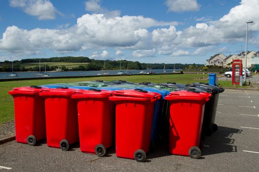 A group of plastic multi-coloured wheelie bins arranged on tarmac infront of a park and a stretch of water with yachts.