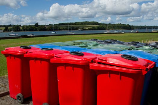 A group of plastic multi-coloured wheelie bins arranged on tarmac infront of a park and a stretch of water with yachts.