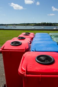 A group of plastic multi-coloured wheelie bins arranged on tarmac infront of a park and a stretch of water with yachts.