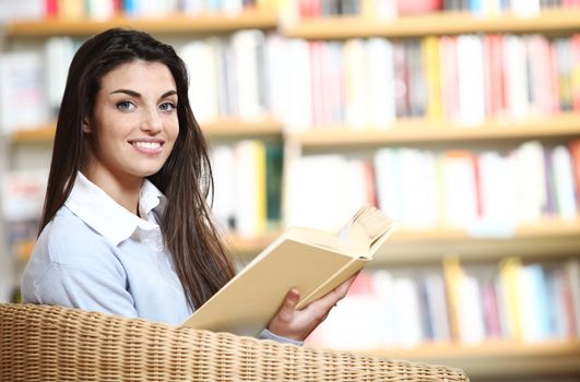 smiling female student with book in hands sitting in a chair in a bookstore - model looking at camera. 