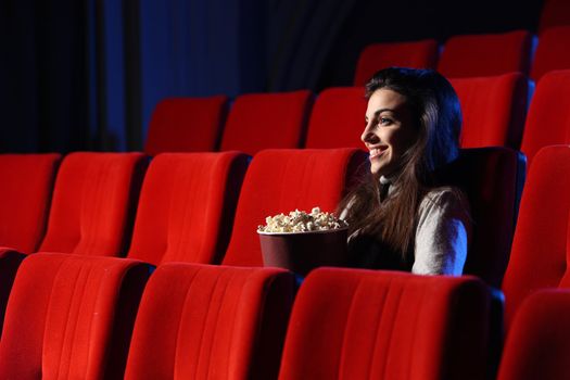 a pretty young woman sitting in an empty theater, she eats popcorn and smiles, side view