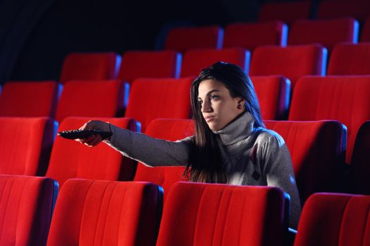 a young woman holding a TV remote control, in the background you can see the red chairs in a movie theater. conceptual image