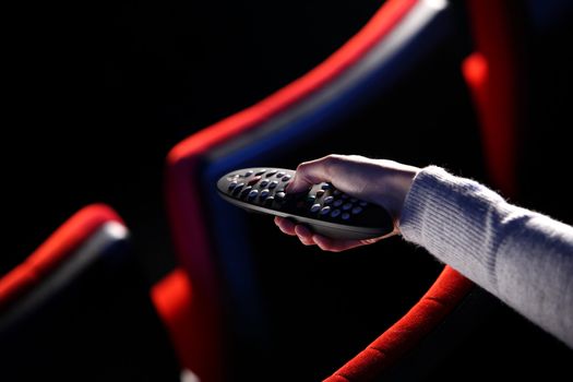 closeup of a hand holding a remote control TV, in the background you can see the red chairs in a movie theater. conceptual image