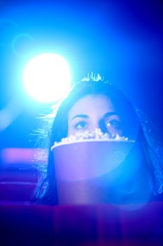 
a pretty young woman sitting at theater, she is scared, and hides behind a box of popcorn