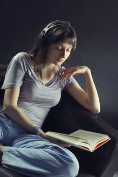 A young woman sits on a couch  reading a book and listening to music through a pair of headphones