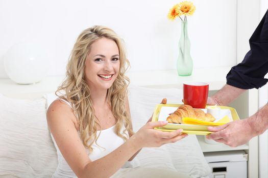 Young woman being treated to breakfast in bed as she is handed a tray with a fresh crisp croissant and coffee