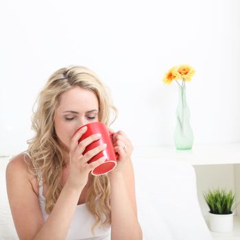 Young woman with long blonde curly hair drinking a large red mug of coffee in bed