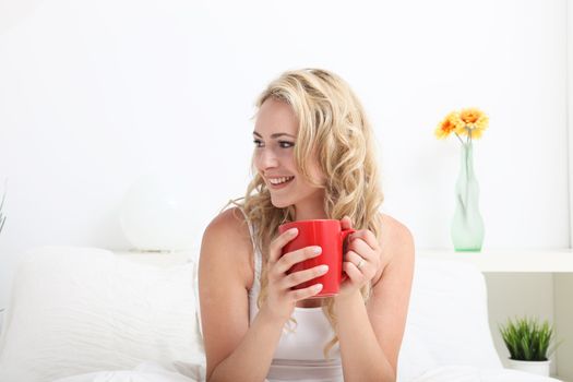 Happy smiling woman sitting in bed drinking coffee and looking sideways off frame across blank copyspace
