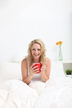 Vivacious young woman with lovely curly blonde hair enjoying a cup of coffee while sitting in bed