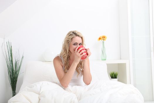 Woman sitting in a comfortable bed with white linen enjoying a bright red mug of coffee