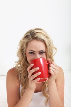 Young blonde woman with smiling eyes drinking a large mug of coffee against a white interior background