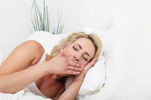 Young woman lying in her comfortable bed yawning with her hand covering her mouth