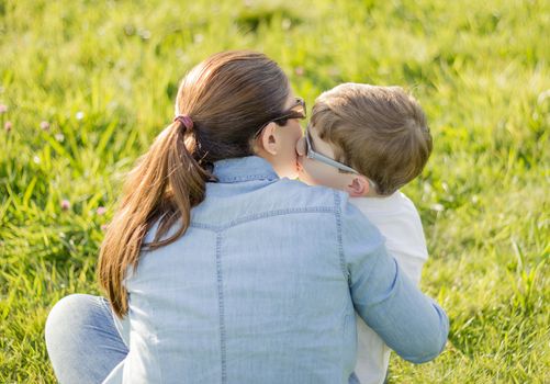 Back view of cute son kissing to his mother sitting in a sunny field