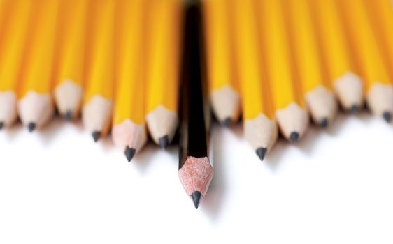 Low angle shot of Uneven row of yellow pencils with one black pencil in middle standing out farther than the rest. Focus is on the tip of the black pencil. On white with drop shadow