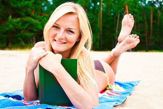 A young and beautiful woman resting on a beach reading a book in hard cover green
