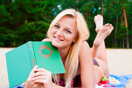 A young and beautiful woman resting on a beach reading a book in hard cover green