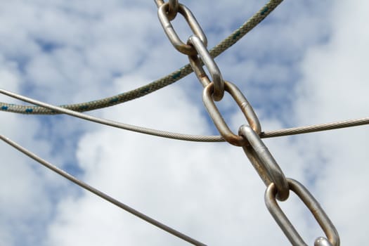 A white metal linked chain with rust lays over a cable and rope against a blue cloud sky.