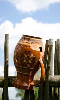 Clay broken pitcher hangs on wooden fence from woven tree branches.